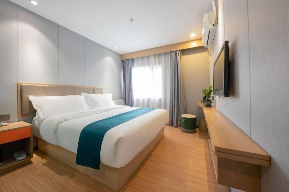 A hotel room with a bed, a bedside table, a small table, a wall-mounted TV, and a window with curtains.