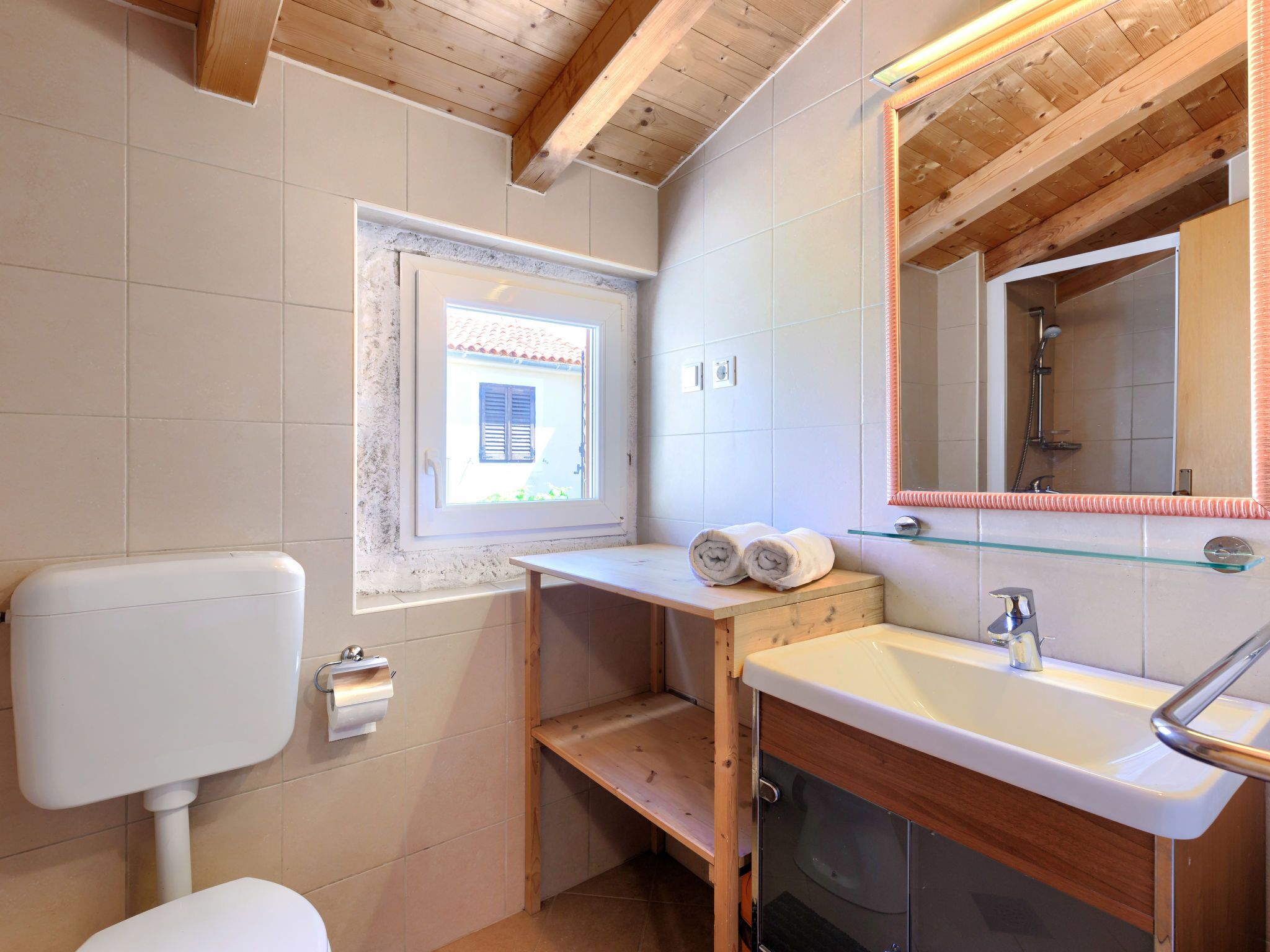 A bathroom with a wooden ceiling, a window with a view, a sink with a mirror, and a toilet.