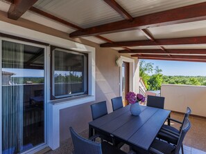A covered patio with a table and chairs, a window with curtains, and a view of greenery.