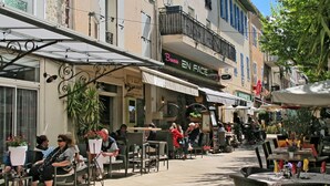 An outdoor café with tables and chairs, surrounded by buildings with various shops and restaurants. - Micocoulier by Interhome (Bagnols en Forêt)