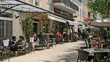 Un café en plein air, avec des tables et des chaises, entouré d’immeubles abritant divers commerces et restaurants.
