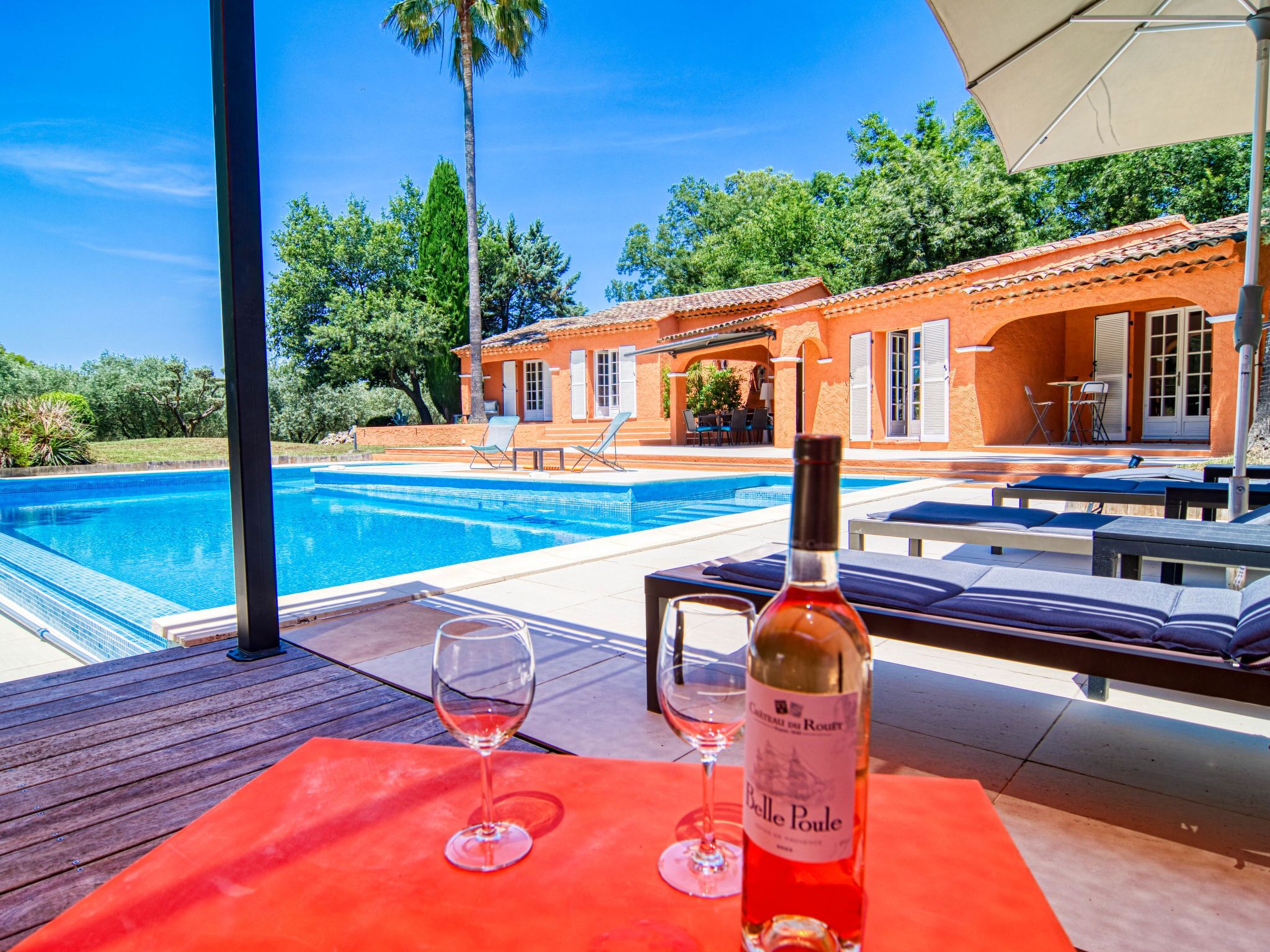 Un espace aménagé au bord de la piscine, avec une terrasse en bois, une bouteille de vin et deux verres.