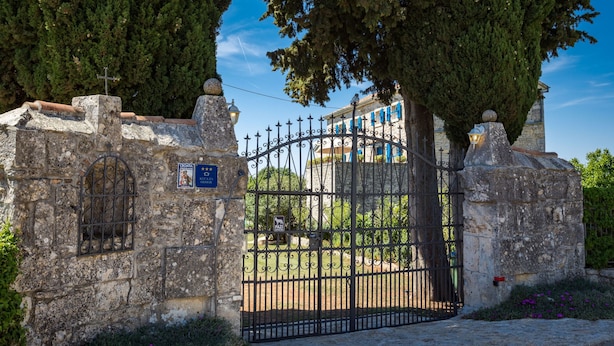 Une porte historique en pierre, surmontée d’une grille métallique, qui mène à un bâtiment aux volets bleus et sur lequel on aperçoit une croix au sommet du mur.