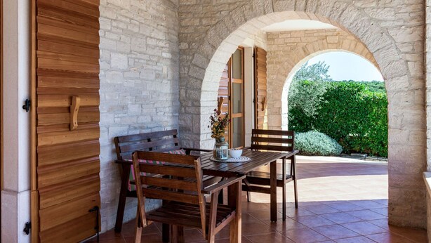 Un patio con una mesa y sillas de madera, un arco y un muro de piedra.