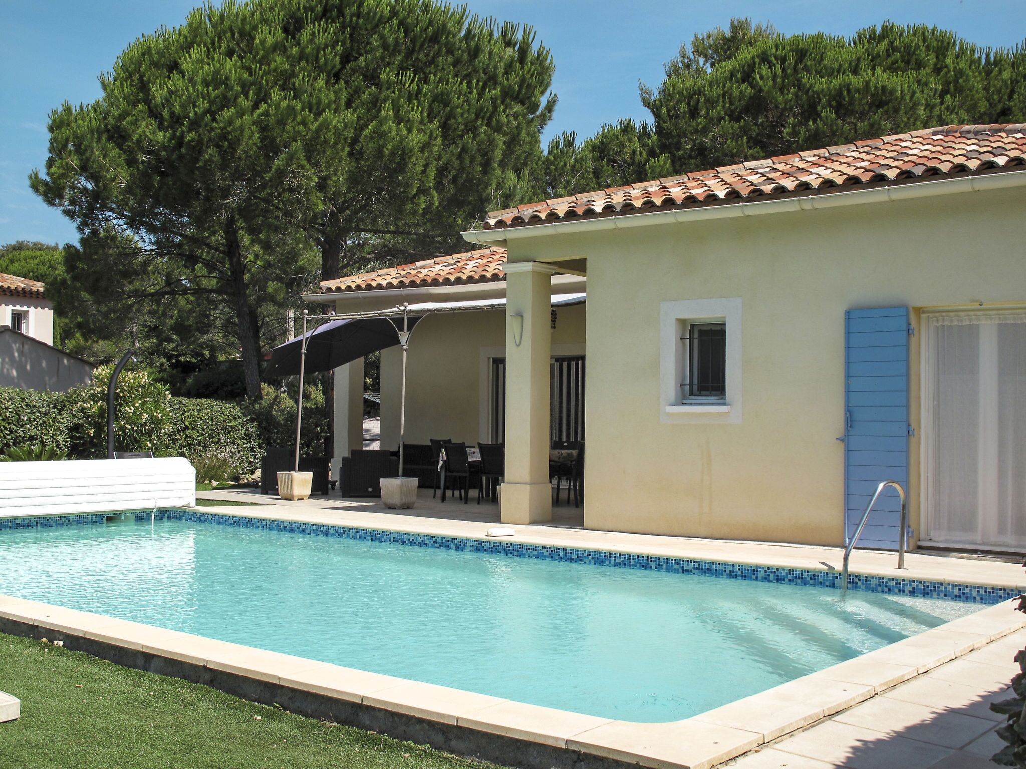 Ein Swimmingpool mit gefliestem Rand, eine überdachte Terrasse mit Gartenmöbeln und ein Gebäude mit einem gepflasterten Dach.