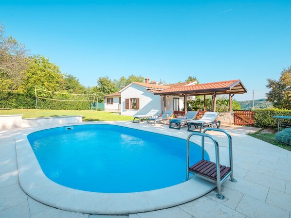 Piscina com espreguiçadeiras e um pergolado, cercada por uma casa e vegetação.
