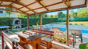 Un patio techado al aire libre con una mesa de piedra, bancos y una piscina.
