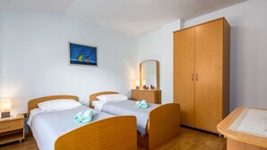 Habitación de hotel con dos camas de madera, un armario grande, un escritorio con silla, una mesita de noche, una lámpara y un cuadro en la pared.