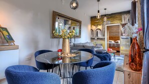 Una sala de estar moderna con una mesa redonda de cristal, sillones de terciopelo azul, un jarrón dorado con flores y un espejo grande en la pared.
