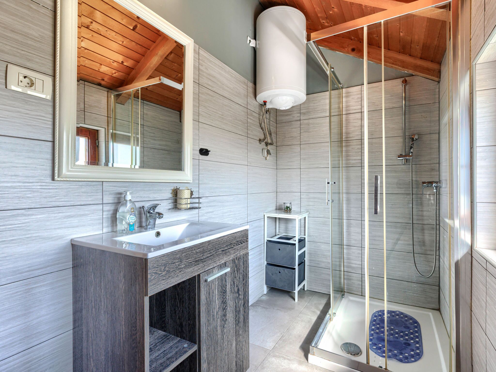 Et moderne badeværelse med træloft, brusekabine i glas, vask med spejl og en lille hylde med toiletartikler.