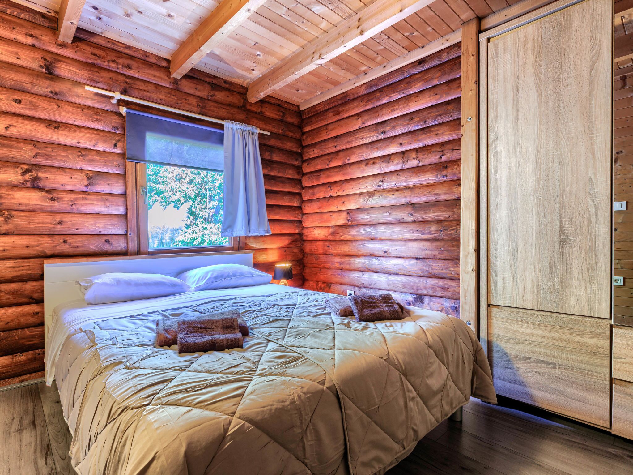 A wooden cabin bedroom with a bed, a window with curtains, and a wardrobe.