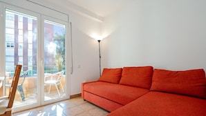 Ein Wohnzimmer mit einem roten Sofa, einem Holzstuhl, einer Stehlampe und einer Schiebeglastür, die zu einem Außenbereich mit Tisch und Stühlen führt.