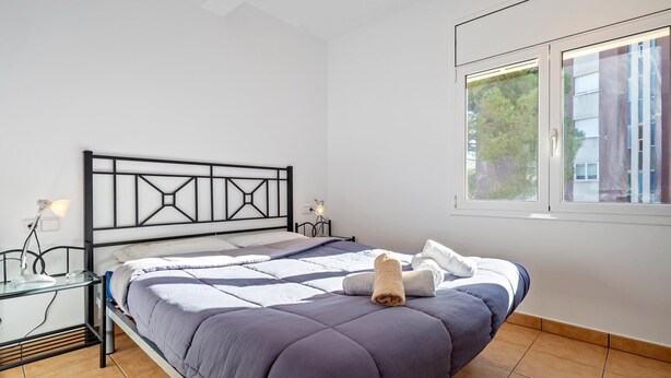 Ein ordentlich bezogenes Bett mit modernem Kopfteil, ein Nachttisch mit Lampe und ein Fenster mit Blick ins Grüne.