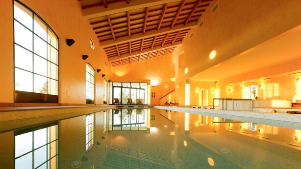 Une grande piscine intérieure à l’eau claire, dotée de poutres en bois au plafond et de grandes fenêtres laissant entrer la lumière naturelle.