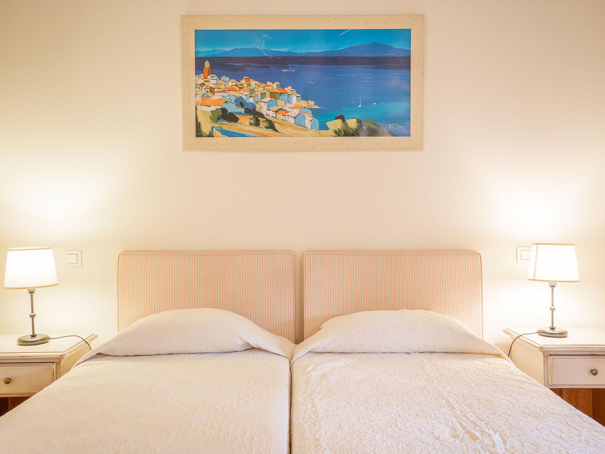 Una cama doble con cabecera a rayas, dos mesitas de noche con lámparas, y un cuadro de un pueblo costero en la pared.