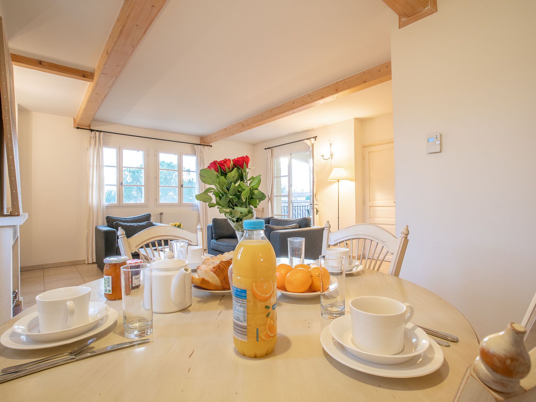 A dining table set with breakfast items, a vase with flowers, and a view of the living area with a sofa and windows.