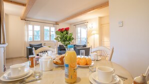 Une table à manger dressée avec des aliments pour le petit-déjeuner, un vase rempli de fleurs et une vue sur le salon, avec un canapé et des fenêtres.