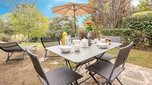 Une terrasse extérieure avec une table dressée pour le repas, entourée de chaises en osier, d’un grand parasol et d’un vase rempli de fleurs.