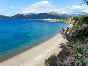Ein Küstenstrand mit klarem, blauem Wasser, sandigem Ufer und Bergen im Hintergrund.