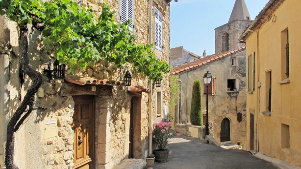 Eine enge Gasse mit Steinhäusern, einem Kirchturm und einem kleinen Innenhof mit einer Bank und Topfpflanzen.