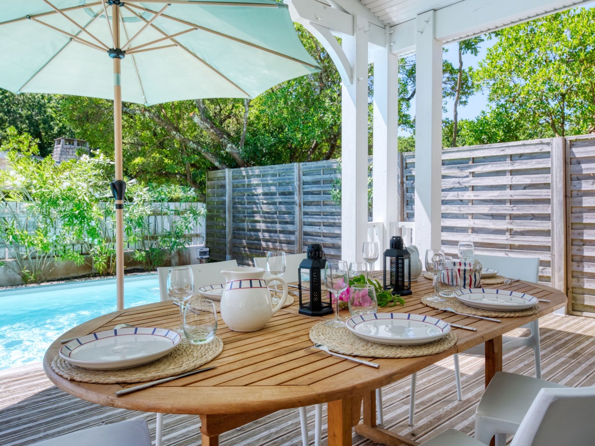 Een terras met een houten tafel gedekt voor vier personen, omringd door witte stoelen, onder een grote parasol.