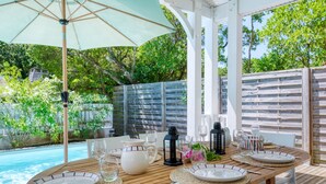 An outdoor dining area with a wooden table set for four, surrounded by white chairs, under a large umbrella. - Vacation home Le Lac by Interhome (Lacanau)