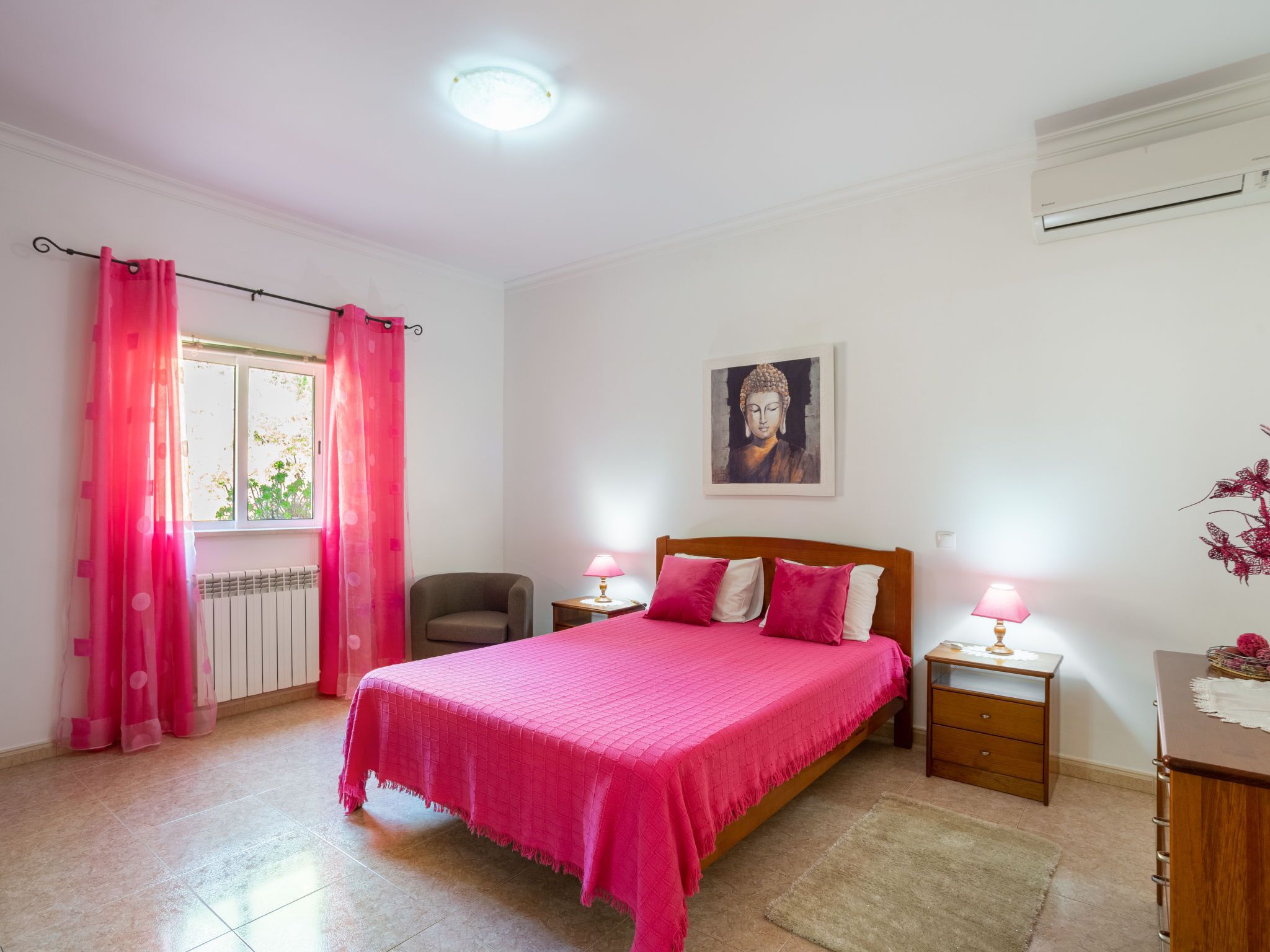 A bedroom with a bed, two bedside tables with lamps, a chair, a radiator, and a window with pink curtains.