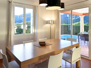 A dining area with a wooden table and white chairs, large windows, and a view of a swimming pool. - Domaine de l'Eilen V68I by Interhome (Cavalaire)