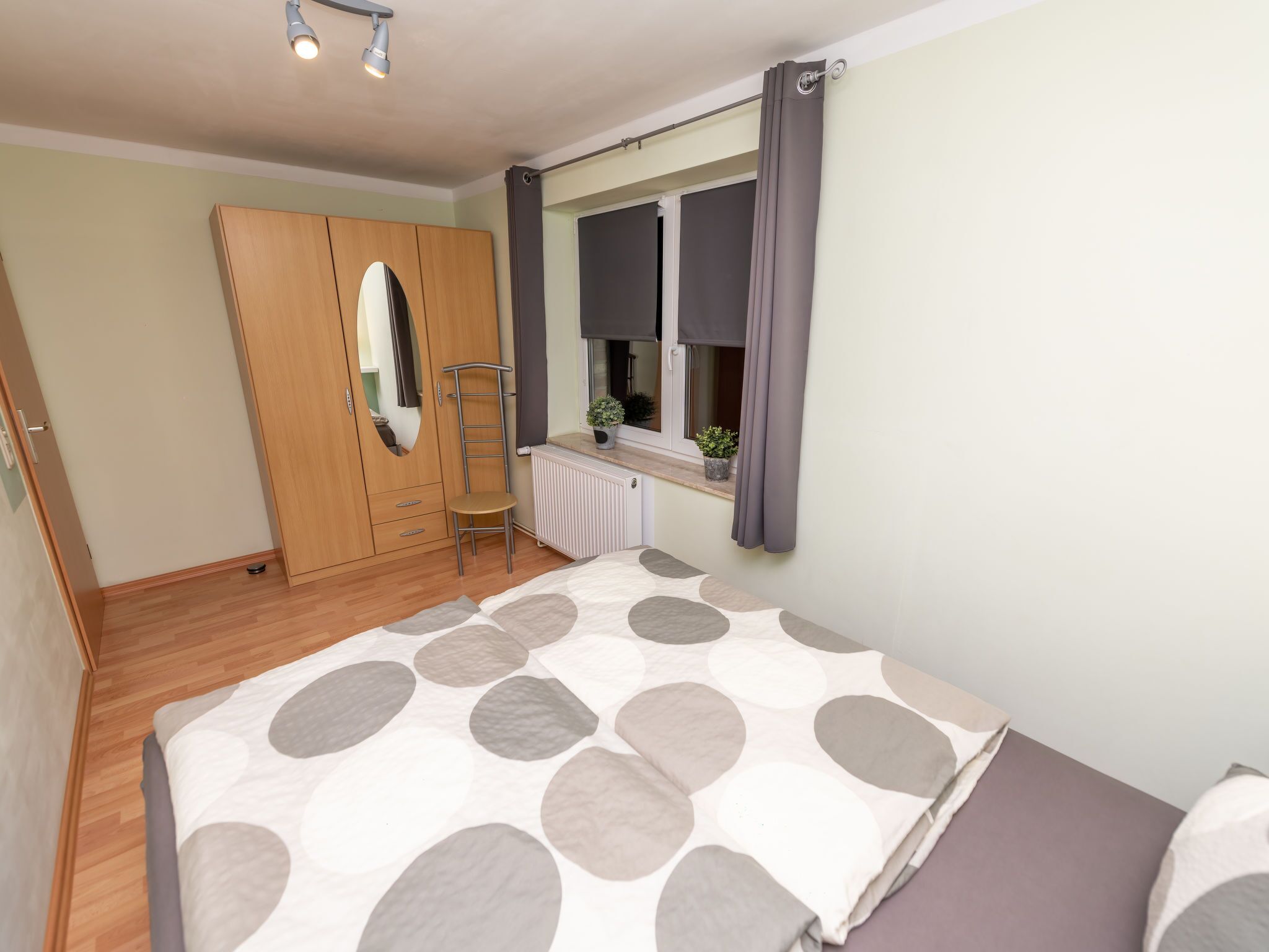 A bedroom with a bed, a wardrobe, a window with curtains, a radiator, and a small plant on the windowsill.