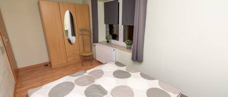 A bedroom with a bed, a wardrobe, a window with curtains, a radiator, and a small plant on the windowsill.