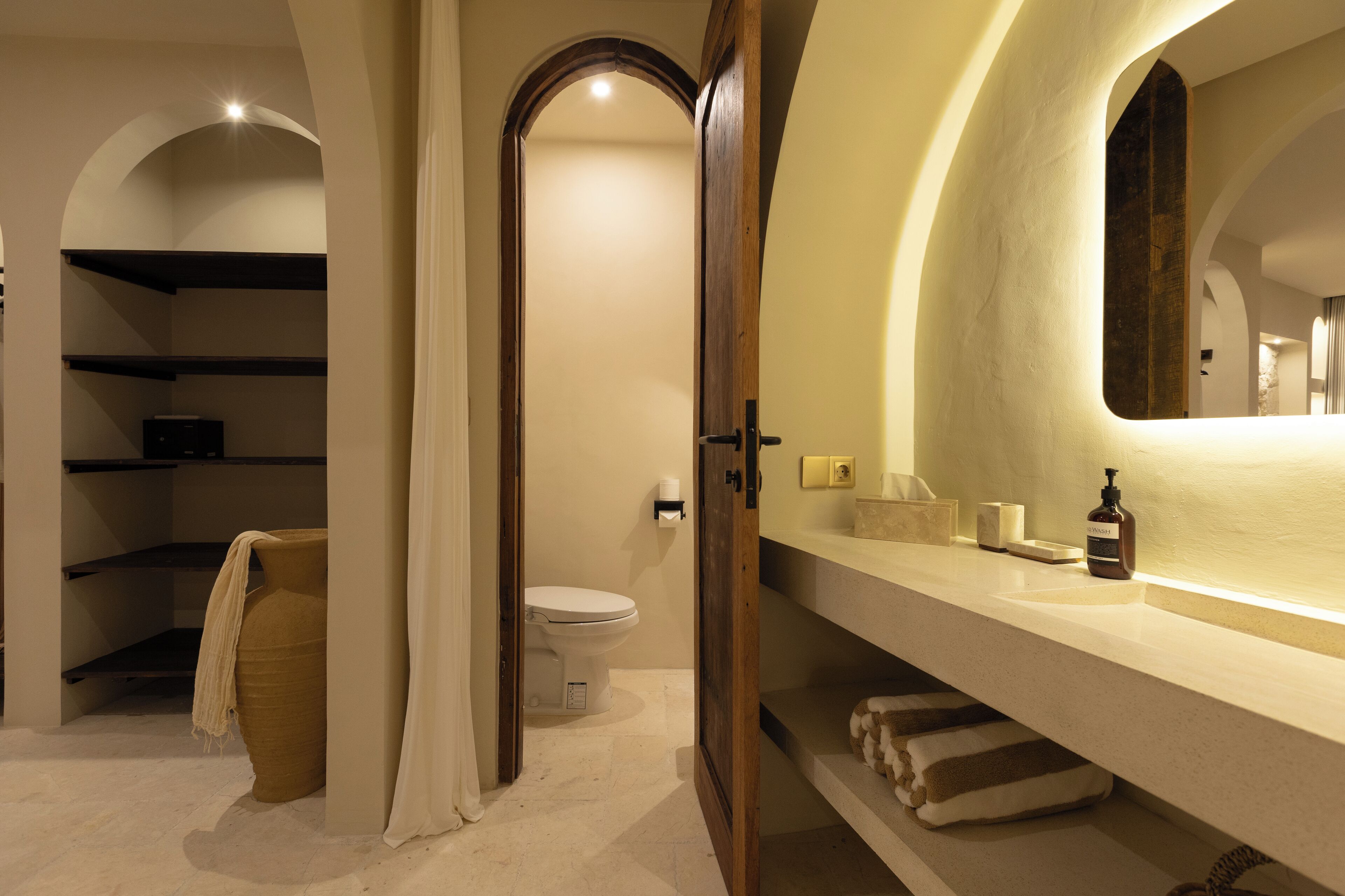 A bathroom with a white sink, a mirror, and a toilet. There are shelves with towels and toiletries.