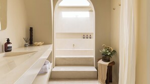 Et badeværelse med et stort spejl, en håndvask med toiletartikler, et bruseområde og en potteplante.