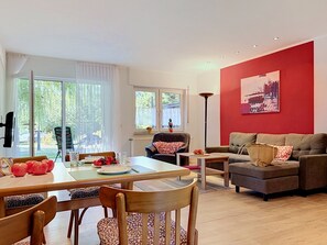 A modern living room with a dining area, a red accent wall, and large windows.