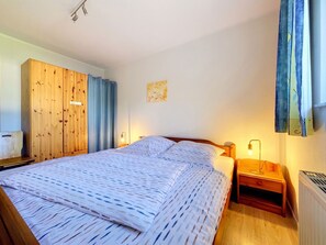 A bedroom with a bed, wooden wardrobe, curtains, a wall art piece, and a bedside table with a lamp. - Lenzer Höh by Interhome (Plau am See)