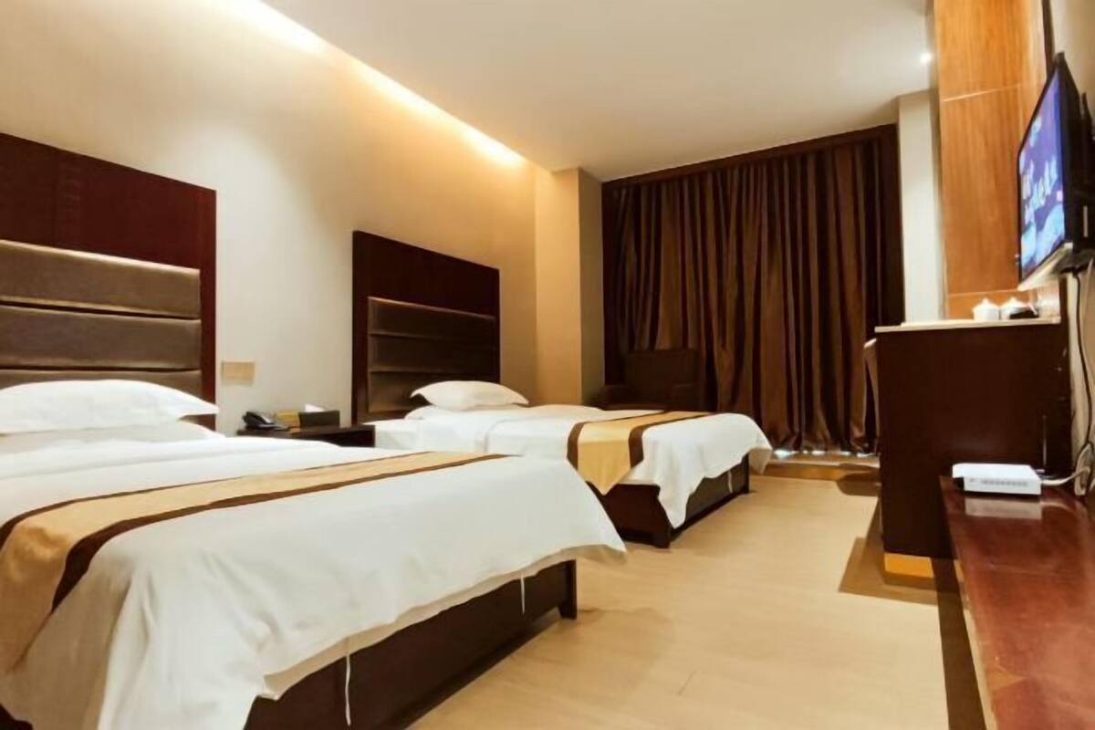 Ein Hotelzimmer mit zwei Betten, einem Fernseher, einem Schreibtisch und einem großen Fenster mit Vorhängen.
