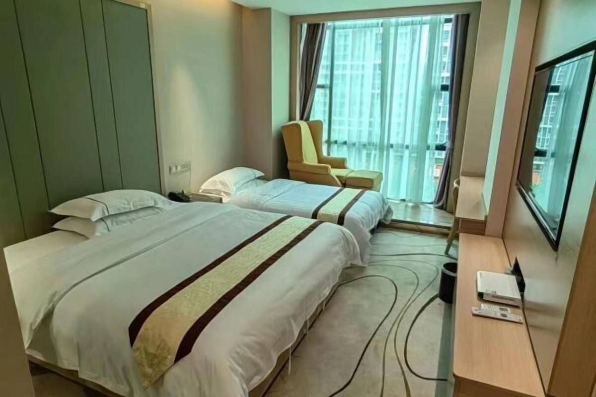 Ein Hotelzimmer mit zwei Betten, einem Sessel, einem Schreibtisch mit Fernseher und einem Fenster mit Vorhängen.