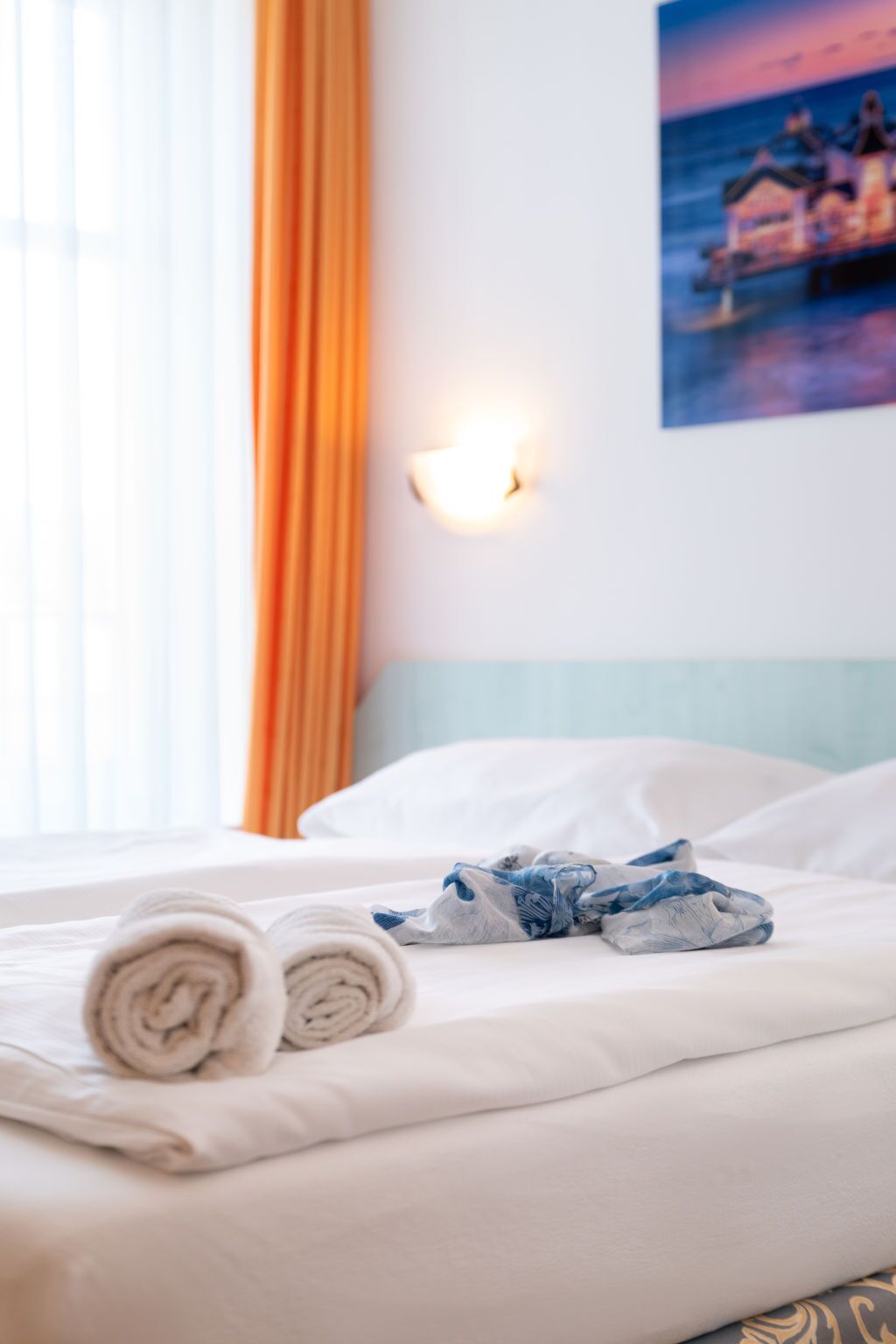 A neatly made bed with rolled towels and a pair of jeans, a wall-mounted light, and a window with curtains.
