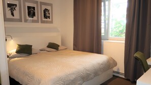 Una cama bien tendida con un edredón blanco texturizado, un cabecero con tres cuadros enmarcados y una ventana con cortinas.