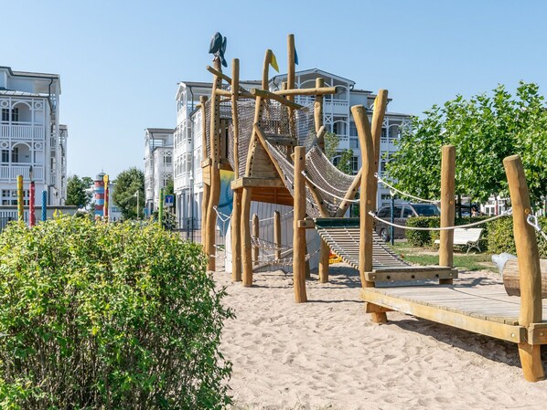 A wooden play structure with a rope bridge and a wooden platform, set against a backdrop of white buildings and a clear sky. - Seepark Sellin by Interhome (Ostseebad Sellin)