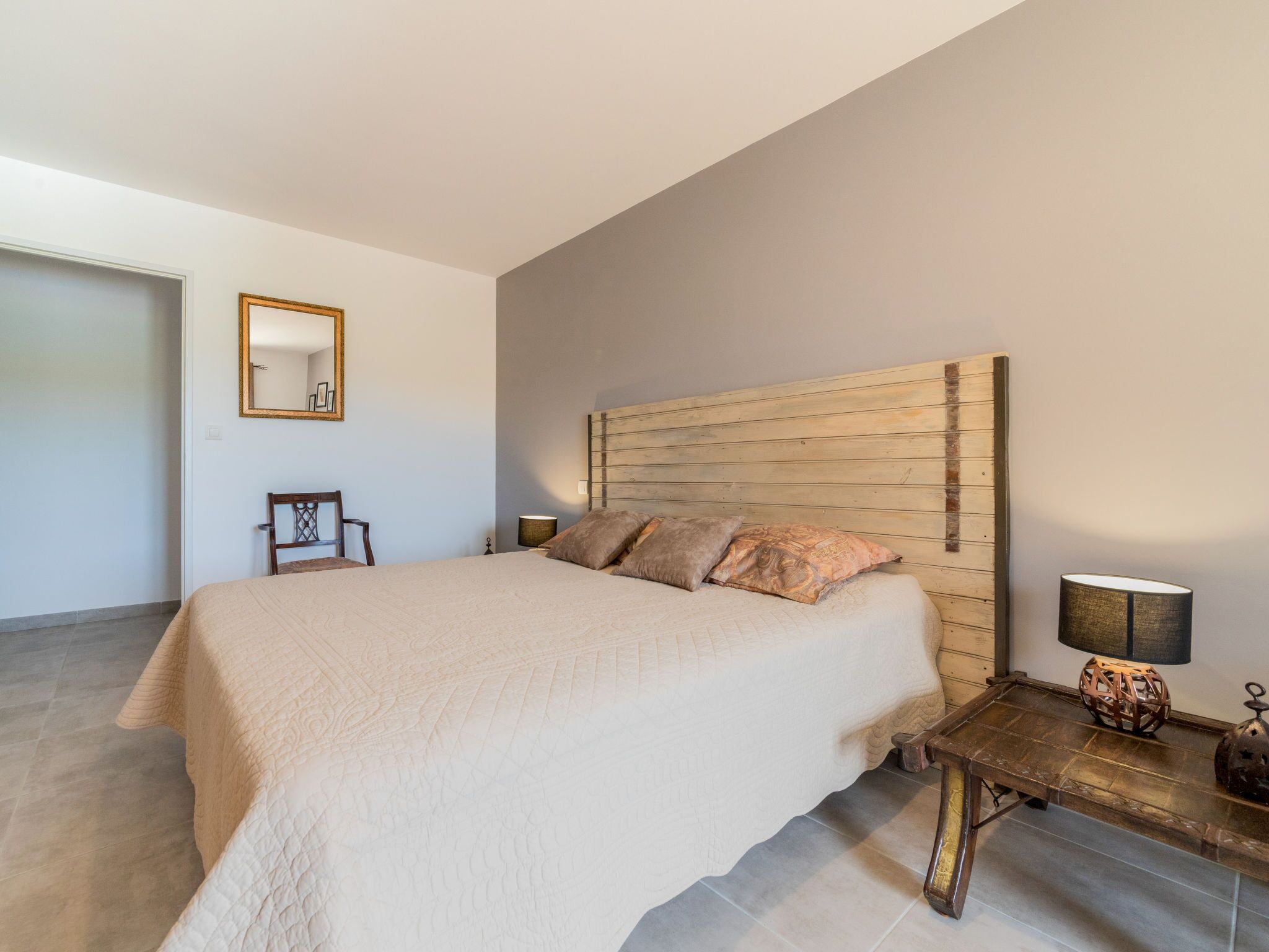 Une chambre à coucher avec un grand lit, une tête de lit en bois, une table de chevet avec une lampe, une chaise et un miroir au mur.