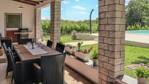 Un patio al aire libre con una mesa de comedor, sillas y una parrilla. Hay plantas en macetas y se ve una piscina y follaje en el fondo.