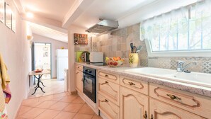 Een keuken met een magnetron, broodrooster en spoelbak. Er is een raam met gordijn en een deur naar een andere kamer.