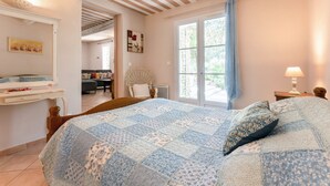 Een slaapkamer met een groot bed, een raam met gordijnen, een nachtkastje met een lamp en uitzicht op de woonkamer.