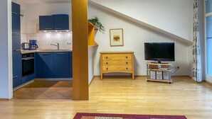 A modern kitchen with blue cabinets, a coffee maker, and a wooden cabinet with drawers. There's a TV on a stand and a plant on a shelf.