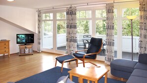 A living room with a sofa, armchair, coffee table, TV, and a balcony with outdoor furniture.
