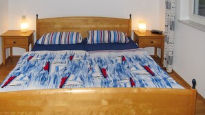 A bed with a blue and white patterned bedspread, two pillows, wooden headboard, two bedside tables with lamps, a small painting on the wall, and a window with a curtain. - Juliusruh by Interhome (Juliusruh)