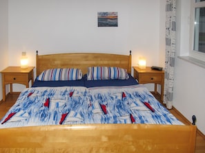 Una cama con colcha de rayas azules y blancas, dos almohadas, cabecera de madera, dos mesitas de noche con lámparas, un cuadro pequeño en la pared y una ventana con cortina.