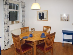 Een eethoek met een ronde houten tafel, vier houten stoelen met rode kussens, een glas water en een blauw kopje.