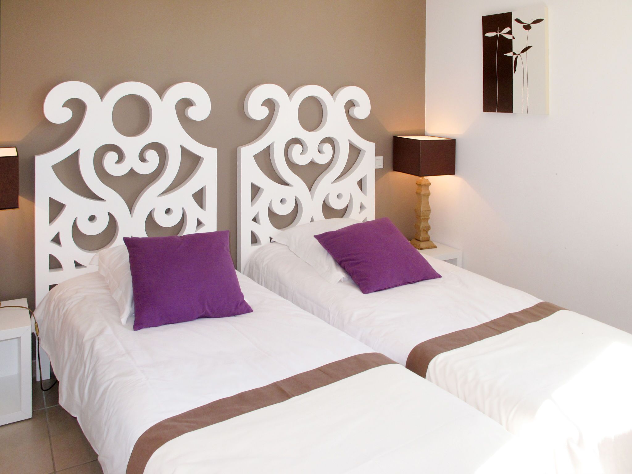 A double bed with white bedding and two purple pillows, a white headboard, a bedside table with a lamp, and a wall-mounted artwork.