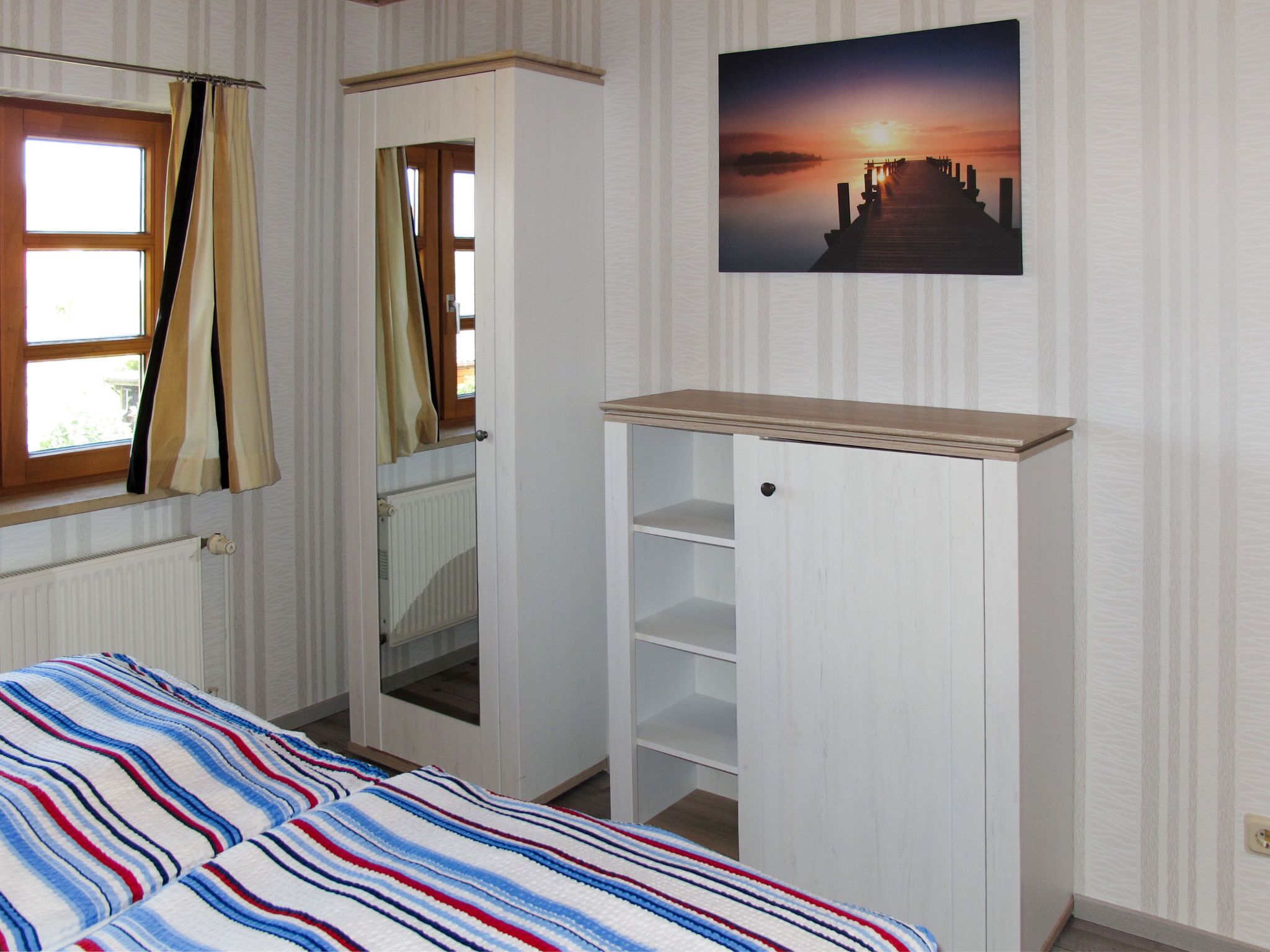Ein Schlafzimmer mit einem Bett, einem Kleiderschrank, einem Heizkörper, einem Fenster mit Vorhängen und einem an der Wand hängenden Bild.